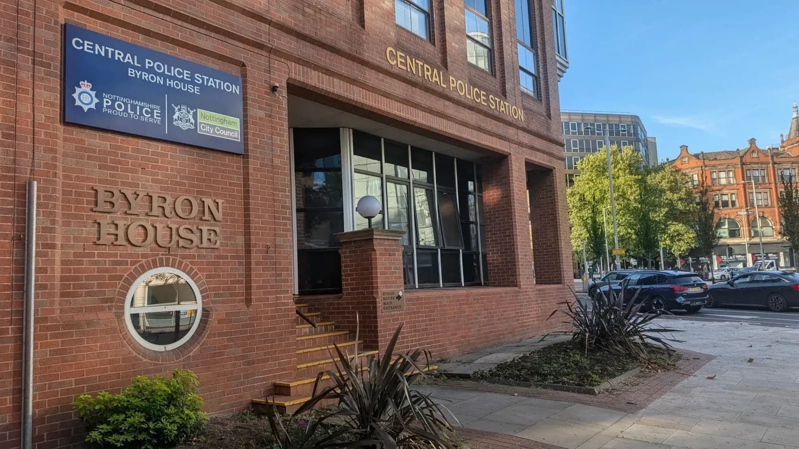 The exterior of the Central Police Station Byron House building with a blue sign on the brick facade and cars parked along the street.