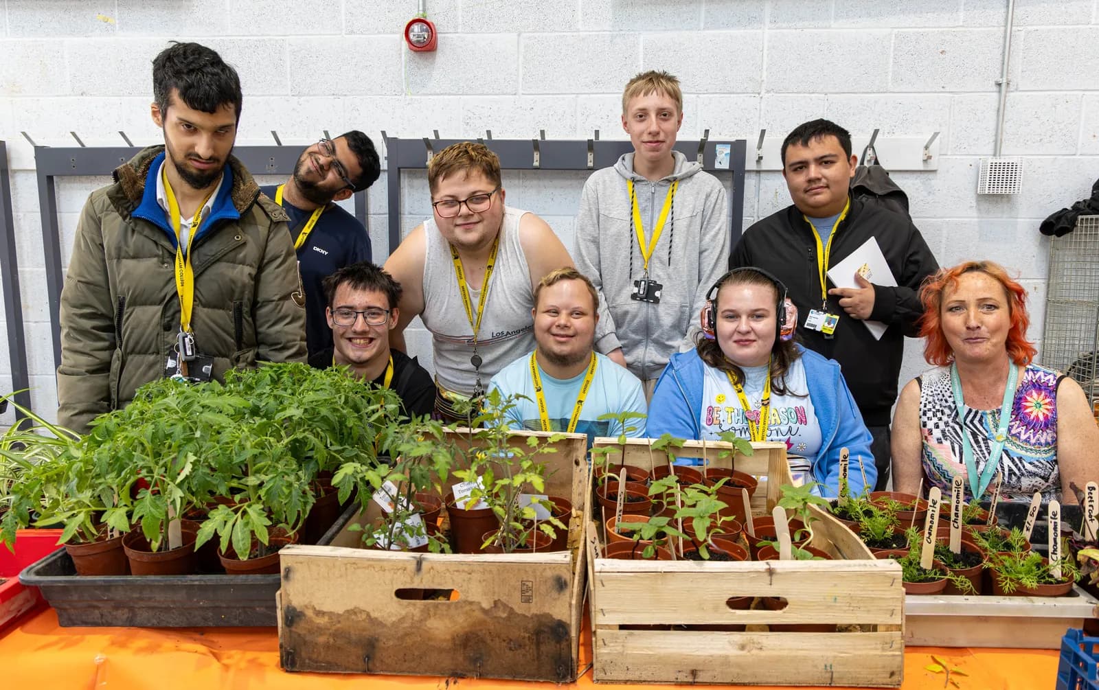A group of eight people, some standing and some sitting, pose behind a table filled with potted plants and seedlings in wooden crates.