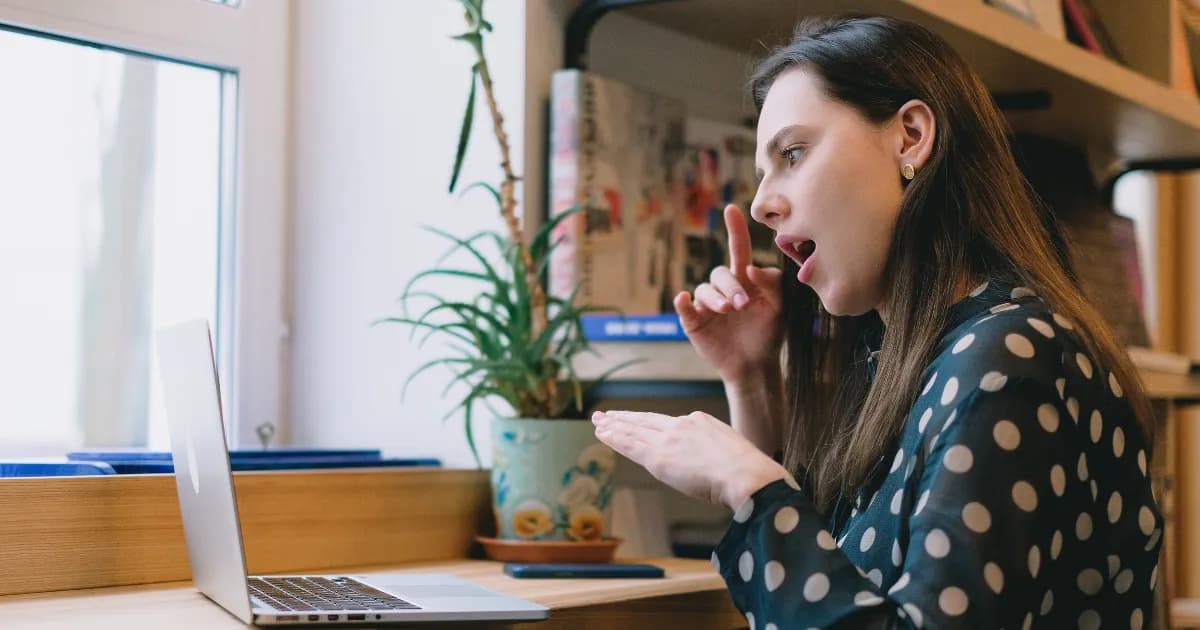 A woman sits at a desk, looking at a laptop and using sign language with her hands.
