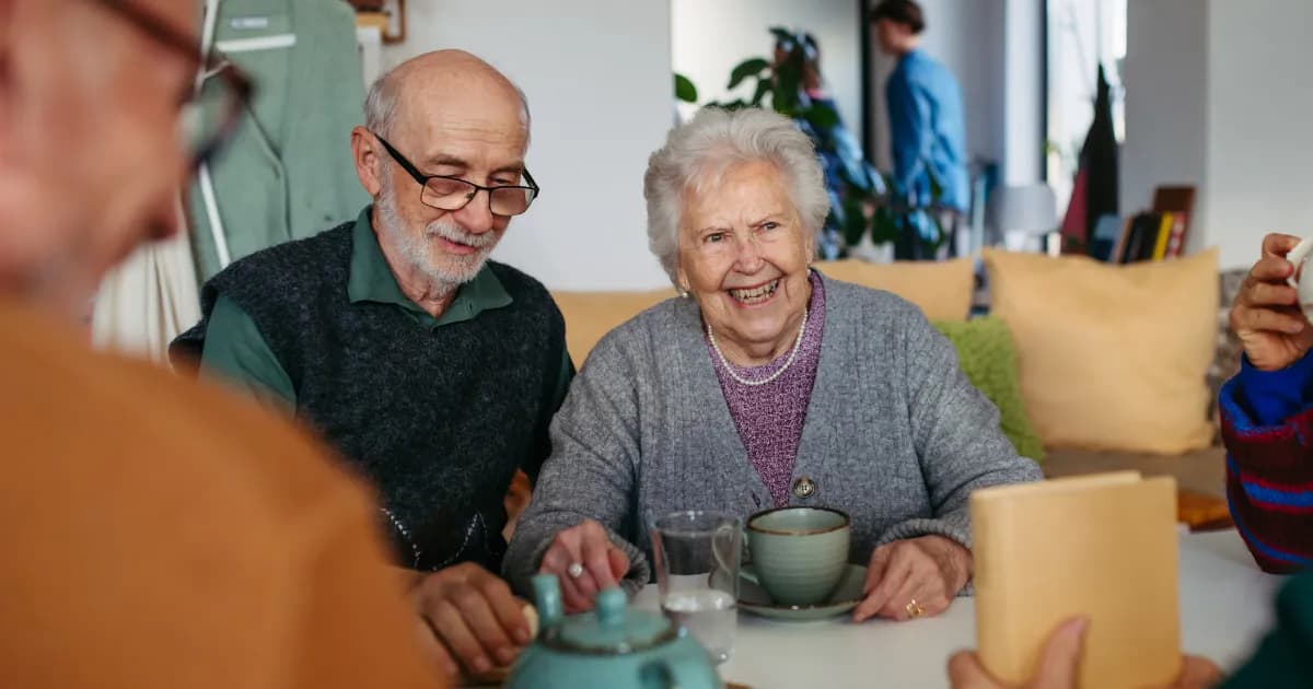 An elderly couple sits at a table, with the woman smiling broadly at the viewer while the man looks down and smiles; another person's blurred back is in the foreground, and a teapot, teacup, and glasses are on the table.