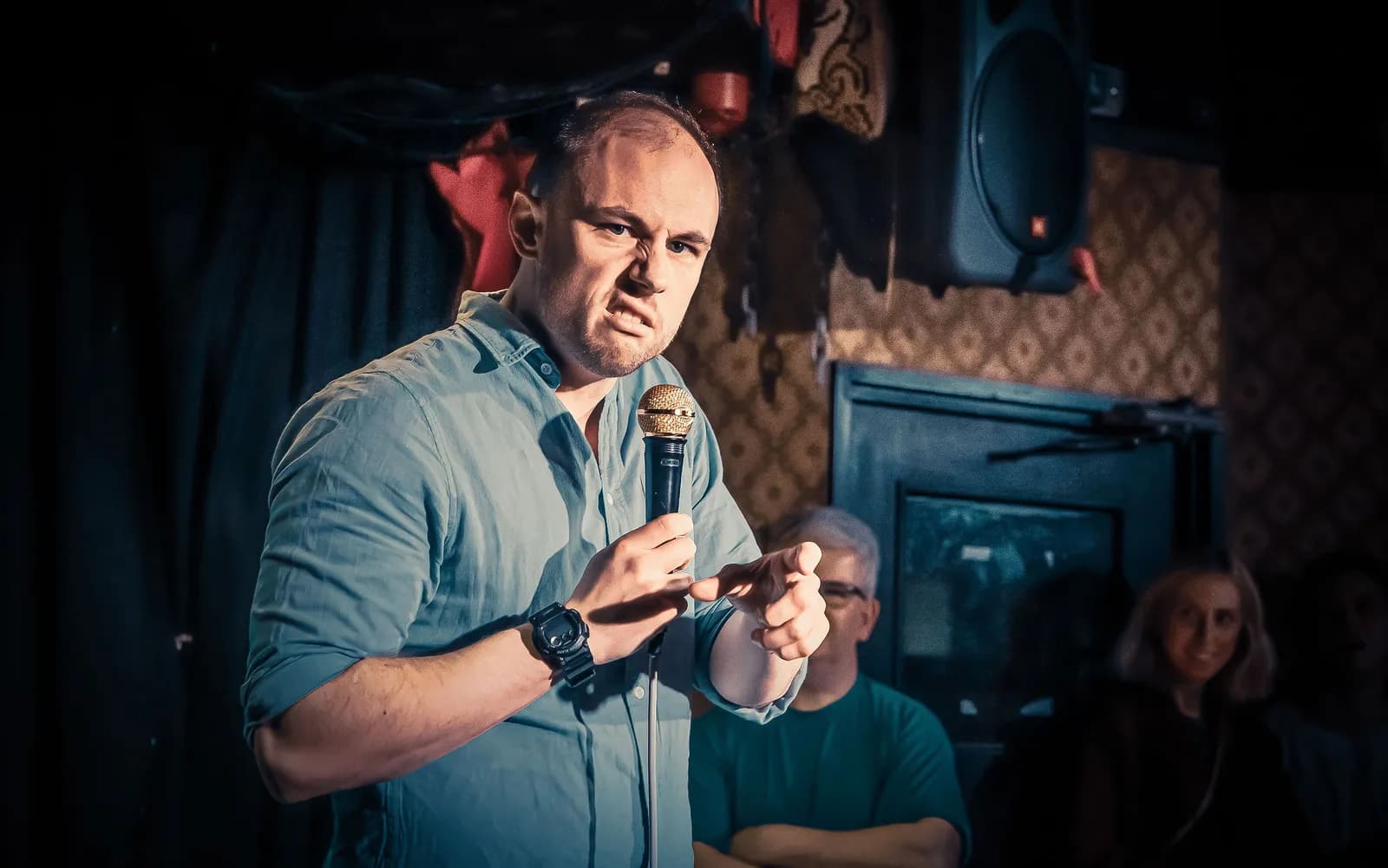 A male comedian with a scrunched-up face holds a golden microphone and points into the audience with his right hand.